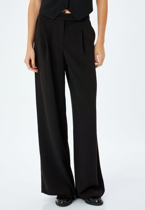 Pantalones anchos negros de tejido suave, con pliegues en la parte delantera y una cintura alta. Combinados con un chaleco negro corto y zapatos planos.