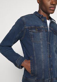 Denim jakna v temno modri barvi z gumbom spričo, dvema žepoma na prsih in gumbi na manšetah. Ima klasičen ovratnik in vidne šive.