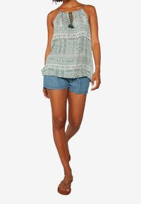 Femme portant un haut dos-nu vert à motifs, un short en jean bleu et des tongs marron, debout avec une jambe croisée sur l'autre.