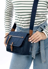 Frau trägt blaue Jeans und ein gestreiftes Langarmshirt, hält eine marineblaue Umhängetasche mit braunen Lederakzenten und silbernem Reißverschluss.