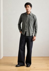 Koszula w paski w oliwkowej zieleni z pojedynczą kieszenią na piersi, w zestawieniu z szerokimi, ciemnoniebieskimi jeansami i czarnymi butami, stojąca na drewnianej podłodze.