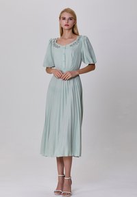 Robe plissée vert menthe clair avec des manches bouffantes, un col rond, des boutons à l'avant et des détails embellis au niveau du col. Sandales blanches à lanières.
