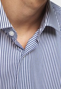 Kraagshirt met smalle blauwe en witte verticale strepen, voorzien van een knoopsluiting en een strakke stoftextuur. Close-up van de halslijn.