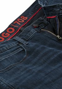 Tmavě modré džíny s rozepínáním na zip, kontrastním červeným vnitřním pasem a raženým logem poblíž pasu. Texturovaná džínová látka.