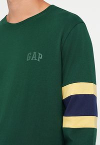Green long-sleeve shirt features GAP logo in light gray. Left sleeve has a yellow and navy stripe pattern. Soft cotton material.