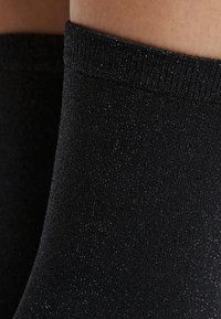 Schwarze glitzernde Knöchelstrümpfe mit einer weichen, strukturierten Oberfläche und einem gerippten Bund, die überall im Stoff einen Hauch von Glanz aufweisen.