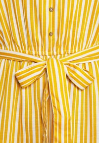 Žlutá a bílá látka se svislými pruhy, s uvázanou mašlí v pase a třemi malými knoflíky nad mašlí.