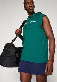 Zelená bezrukávová mikina s logem v bílé a červené barvě, spárovaná s námořnickými šortkami. Model drží černou sportovní tašku. Hladká textura.