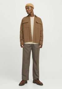 Brown button-up shirt with two chest pockets over a light sweater, paired with grey trousers and brown shoes, topped with a beige beanie.