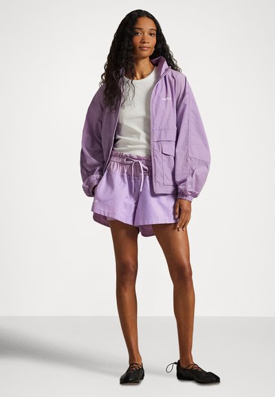 Νέα γυναίκα με μωβ αδιάβροχο μπουφάν και matching σορτσάκι, λευκή μπλούζα και μαύρα δερμάτινα παπούτσια με κορδόνια, στέκεται μπροστά σε λευκό φόντο.