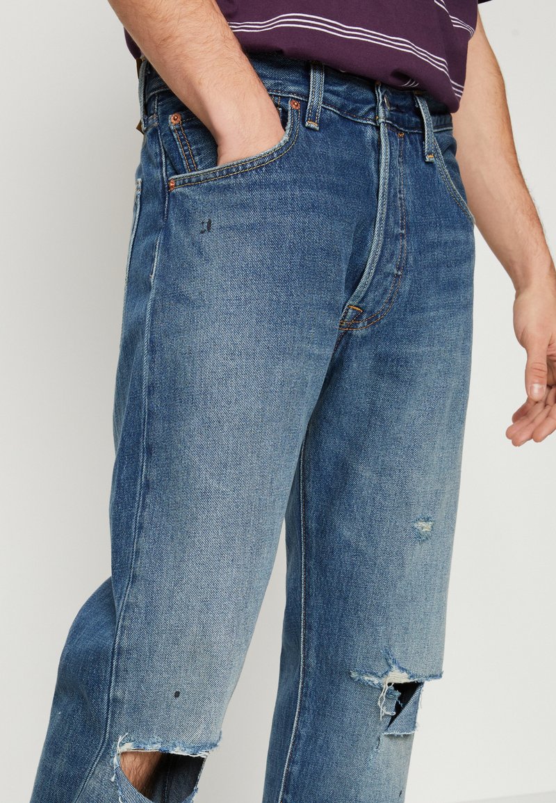 Jean en denim bleu avec une coupe décontractée, présentant de grandes déchirures aux deux genoux, un léger délavage et des accents en métal cuivré.