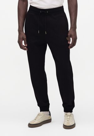 Homme portant un pantalon de jogging noir avec une taille à cordon et des baskets beiges, debout devant un fond blanc uni.