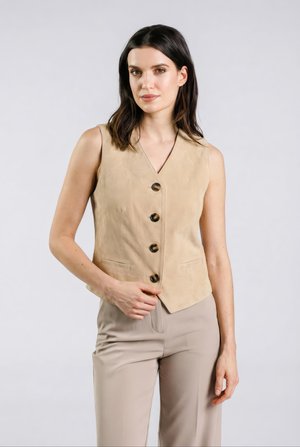 Kvinde iført en beige ærmeløs vest med knapper og lyse taupebukser, står mod en ensfarvet lys baggrund.