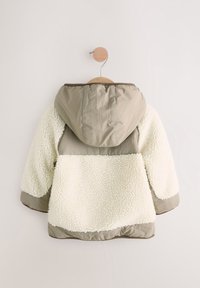 Veste à capuche pour enfant avec tissu en nylon beige sur le dessus et les manches, et polaire sherpa blanche sur le corps et le bas des manches, suspendue à un crochet en bois.