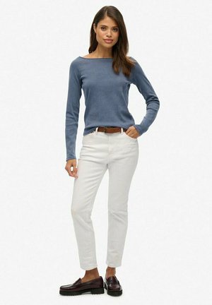 Vrouw staand, gekleed in een blauw shirt met lange mouwen, witte broek met een bruine riem en donkerbruine loafers, tegen een effen witte achtergrond.