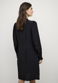 Zwarte jurk met lange, opgeblazen mouwen, gerimpelde manchetten en een relaxte silhouet. Beschikt over een achterkantplooi voor extra bewegingsvrijheid. Zachte stoftextuur.