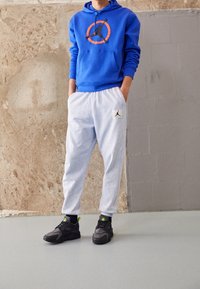Blauer Hoodie mit schwarzer und orangener Grafik, hellgraue Jogginghose mit Jordan-Logo und schwarze Sneakers mit neonfarbenen Akzenten. Texturierter Hintergrund.