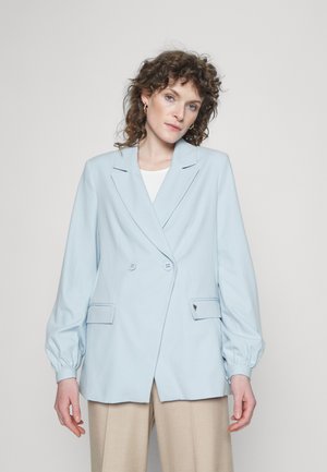 Blazer azul-claro de dois botões em tecido suave com mangas bufantes, apresentando dois bolsos frontais e um emblema discreto no lado esquerdo.