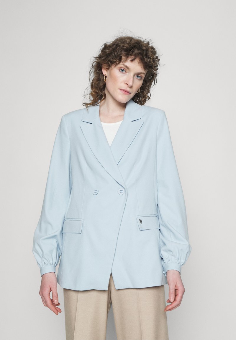 Blazer azul-claro de dois botões em tecido suave com mangas bufantes, apresentando dois bolsos frontais e um emblema discreto no lado esquerdo.