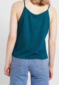 Turkos ärmlös topp med justerbara axelband; mjuk, slät textur; något lös passform; matchad med ljusblå hängande jeans.