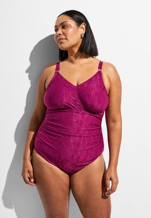 Kvinde med skulderlangt hår iført en struktureret magenta heldragt, stående mod en hvid baggrund, kigger mod højre.