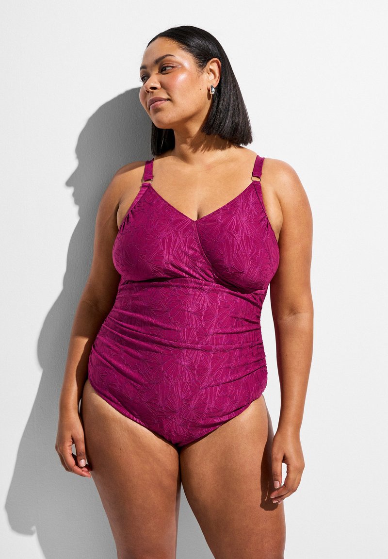 Femme aux cheveux mi-longs portant un maillot de bain une pièce texturé magenta, debout devant un fond blanc, regardant vers la droite.