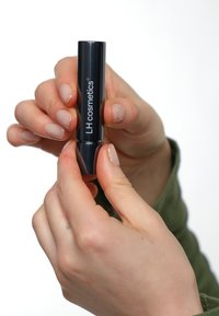 Svart leppestift tube holdt mellom fingrene, med teksten "LH cosmetics." Overflaten er glatt med en blank finish og en avsmalnet tupp.