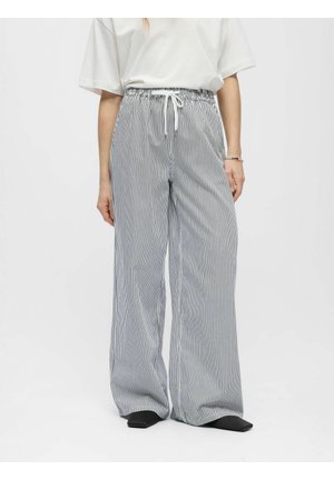 Femme portant un pantalon ample, taille haute, à rayures verticales noires et blanches avec un cordon blanc et des chaussures noires pointues.