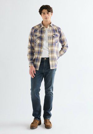 Jonge man die een beige en blauw geruit overhemd draagt over een wit T-shirt, donkere jeans en bruine schoenen, staand tegen een effen witte achtergrond.