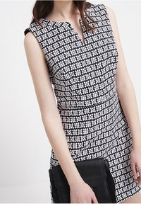Mustavalkoinen kuvioitu ilmanhiha dressi, jossa on V-pääntie ja vartalonmyötäinen leikkaus. Mustaa clutch-laukkuua pidetään vasemmassa kädessä.