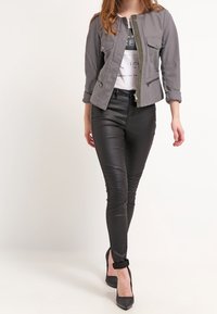 Chaqueta gris corta con dos bolsillos frontales y cierre de botones, combinada con pantalones de imitación cuero negros con detalles acanalados y tacones negros.