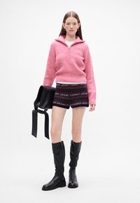 Roza pletena majica s polu-zipom, šarene kratke hlače s natpisom "TOMMY JEANS" i crne kožne čizme do koljena. Crna torba u ruci.