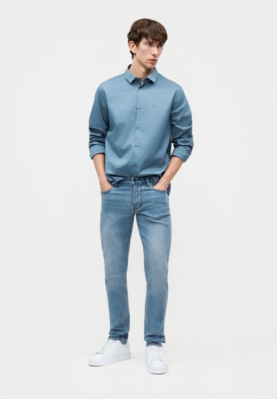 Άνδρας που φοράει ανοιχτό μπλε πουκάμισο με κουμπιά, με τα μανίκια σηκωμένα, ανοιχτό μπλε τζιν, λευκά αθλητικά παπούτσια, στέκεται με τα χέρια στις τσέπες.