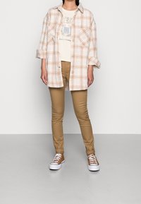 Kostkovaná košile v krémové barvě s oranžovými pruhy, nošená přes světle béžové tričko s potiskem, v kombinaci s těsnými tan kalhotami a hnědými teniskami.