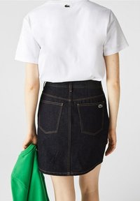 Džínová sukně s tmavým omytem, zdobená oranžovým šitím, má dvě zadní kapsy. Bílá košile a zelená mikina jsou přehozeny přes ruku.