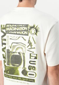 T-shirt en coton blanc avec un design graphique comprenant du texte vert et noir, des formes abstraites et une image d'une bouche, le tout agrémenté d'un motif de soleil ludique.