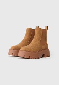 Botas de ante marrón hasta el tobillo con suelas de goma gruesas y paneles laterales elásticos sobre un fondo blanco liso.
