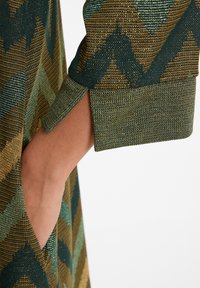 Tissu tricoté avec un motif géométrique en vert, or et marron. Présente un poignet replié avec une poche visible sur le côté.