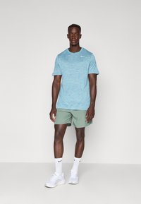 Lyseblå treningsskjorte med strukturert mønster, kombinert med grønne shorts. Hvite joggesko og sokker fullfører det sporty utseendet. Nike-logoen er synlig.