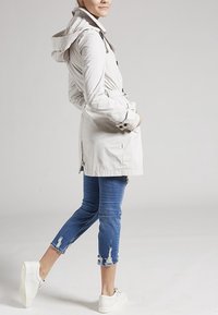 Parka impermeable de un beige claro, con capucha, cinturón ajustable y puños con botones, combinada con unos vaqueros azul desgastados y zapatillas blancas.