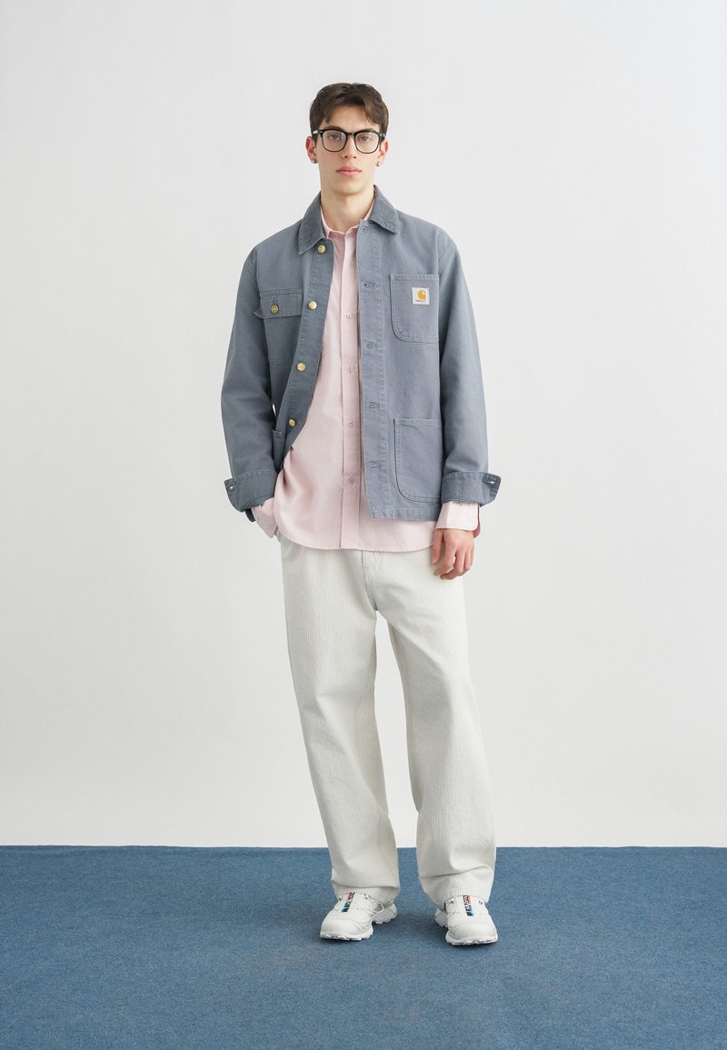 Giovane uomo con occhiali, giacca grigia abbottonata sopra una camicia rosa, pantaloni bianchi e sneakers bianchi, in piedi su un tappeto blu con sfondo bianco.