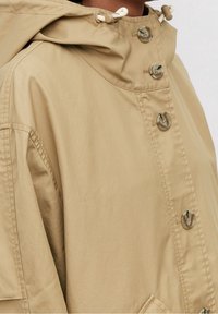 Chaqueta ligera beige con capucha ajustable, cierre de botón y bolsillos laterales. Textura suave y brillo sutil.