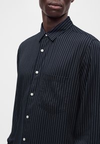 Homme portant une chemise bleu marine foncé à fines rayures blanches verticales et boutons blancs, montrant le col et la poche poitrine.