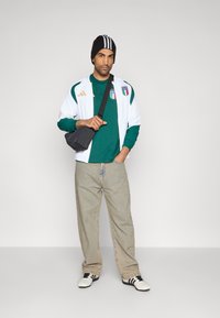 Hvid atletisk jakke med grønne detaljer, grøn langærmet skjorte, lyseblå cowboybukser, sort-hvid beanie og sort skuldertaske.