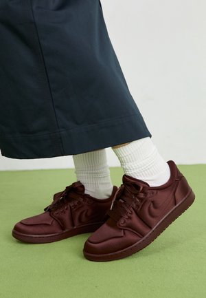 Persona que lleva zapatillas Nike marrón oscuro, calcetines blancos acanalados y pantalones anchos azul oscuro de pierna ancha, de pie sobre un suelo verde.