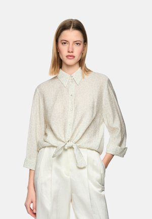 Young woman wearing a light patterned button-up shirt tied at the waist and high-waisted white pants, standing with one hand in pocket.