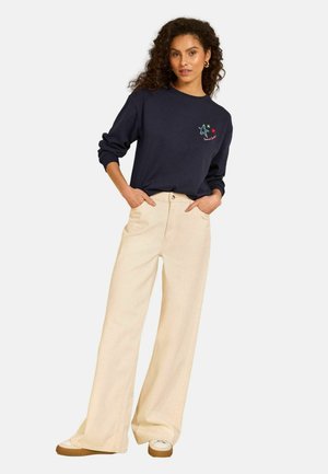 Lokkis juustega naine, kes kannab tumelõhelist pusa tähekujundustega ja beežikaid laiade säärega pükse, seisab käed taskus.