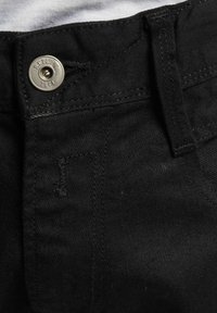 Nærbilde av svart denim jeans midjebånd med sølvfarget knapp graverte "REPLAY" og beltestropp på hvit bakgrunn.