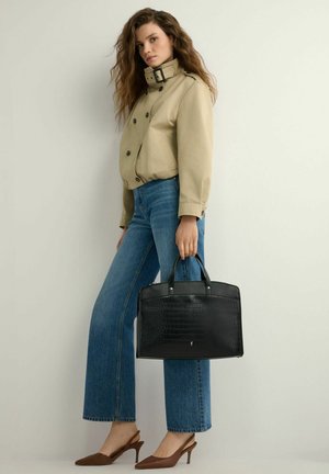 Kobieta o falowanych włosach, ubrana w beżową kurtkę i niebieskie dżinsy, trzymająca czarną teksturowaną torebkę, stojąca na tle jednolitego jasnego tła.