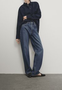 Jean en denim bleu foncé à coupe décontractée, avec une taille haute, un design classique à cinq poches et des détails de couture subtils, associé à une chemise marine.